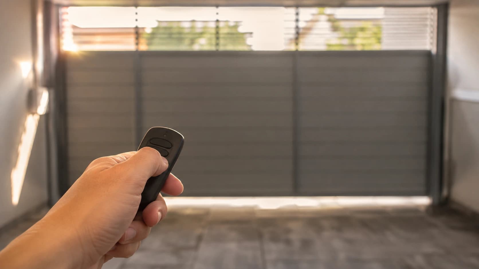 Mão apontando um controle remoto para um portão de garagem de alumínio fechado, ao entardecer