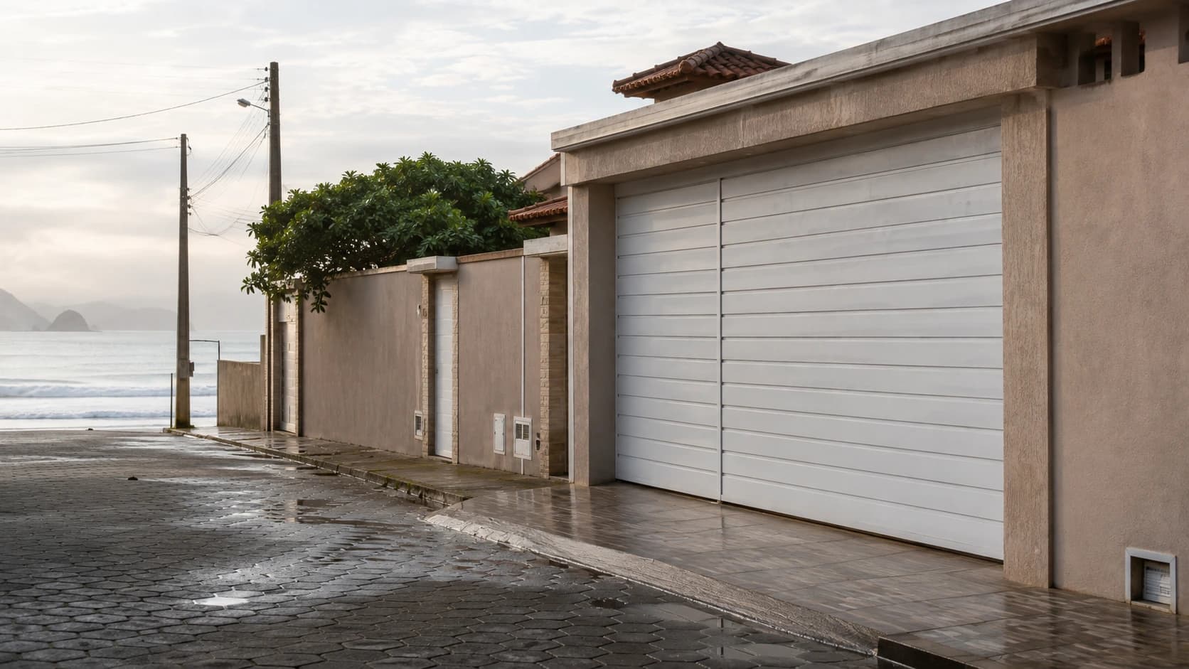 Casa de rua litorânea com portão claro de alumínio fechado e o mar visível ao fundo pela manhã