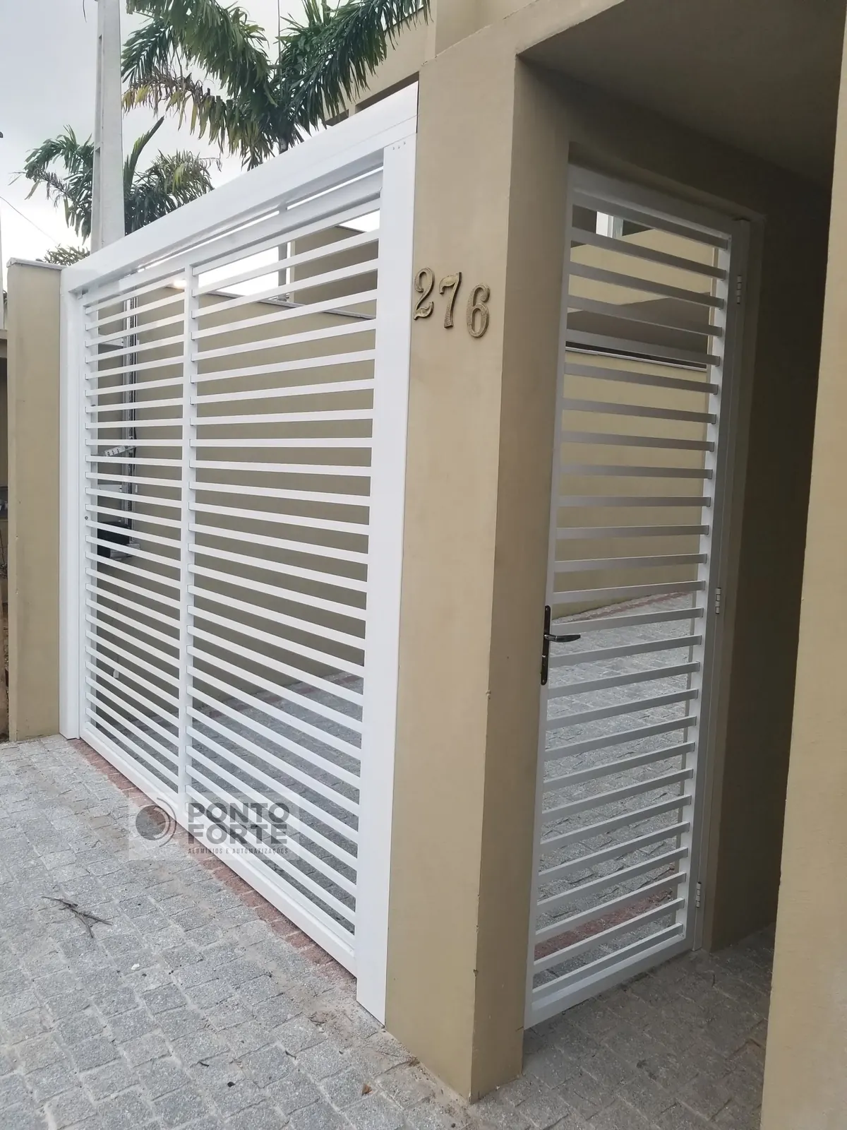 Portão basculante de alumínio branco vazado com barras horizontais, com portão social em vão próprio ao lado, fabricado e instalado pela Ponto Forte