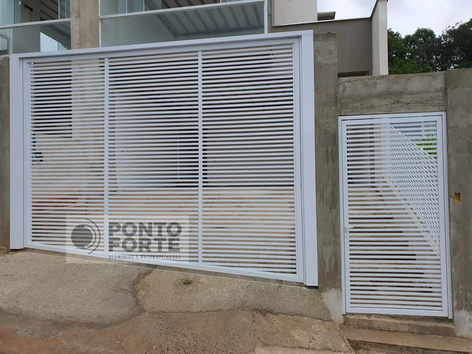 Portão basculante de alumínio branco vazado com barras horizontais, com portão social em vão próprio ao lado, em residência, em Joinville