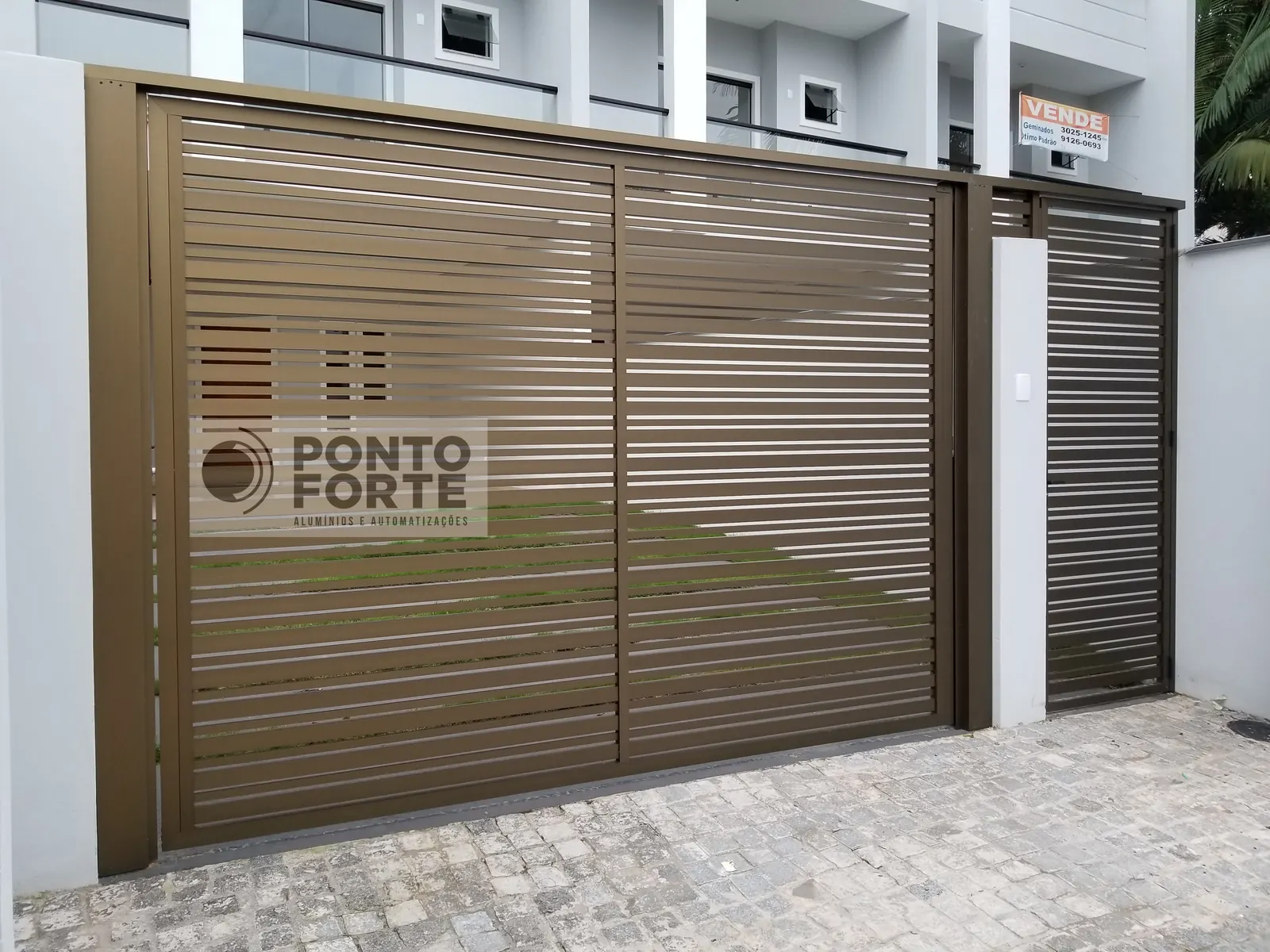 Portão basculante de alumínio bronze vazado com barras horizontais, com portão social em vão próprio ao lado, fabricado e instalado pela Ponto Forte