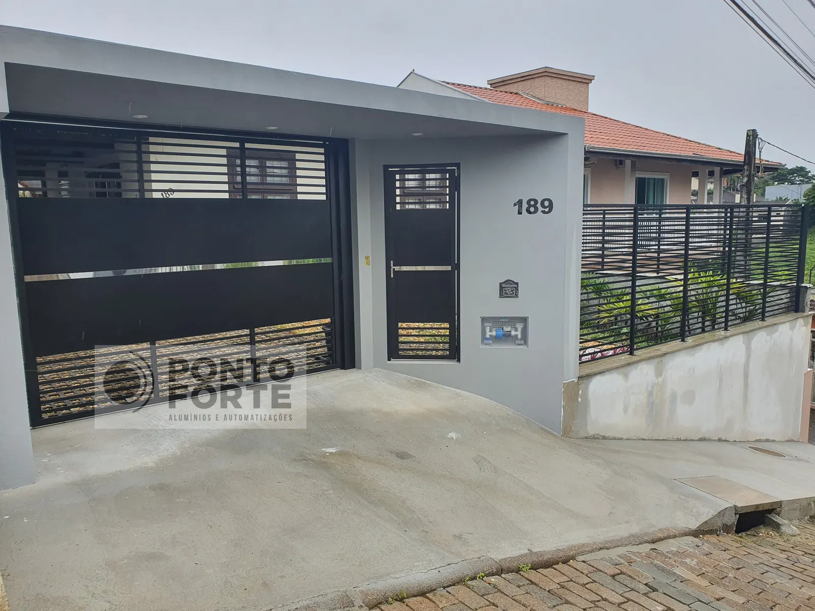 Portão basculante de alumínio preto fechado no centro e vazado em cima e embaixo, com portão social em vão próprio ao lado, em residência, em Joinville