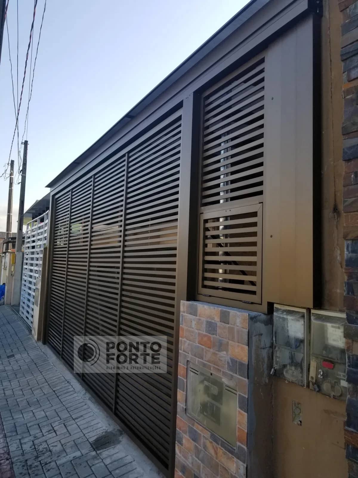 Portão basculante de alumínio bronze vazado com barras, com portão social em vão próprio ao lado, modelo que fabricamos e instalamos em Navegantes