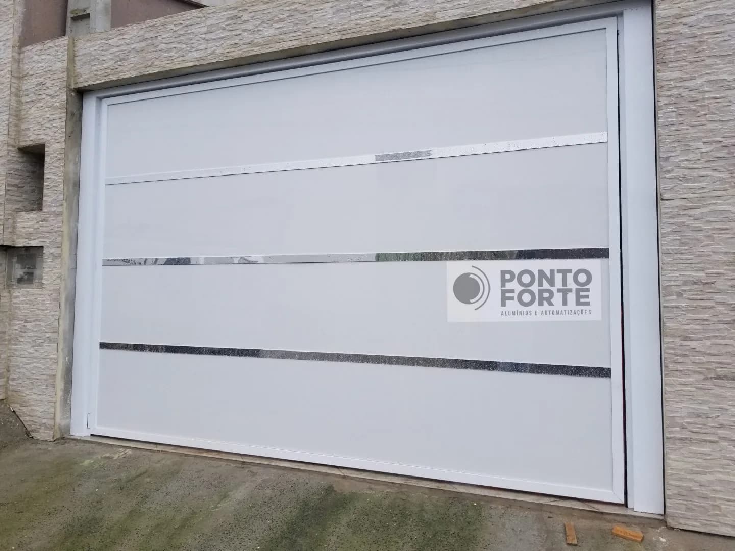 Portão basculante de alumínio branco de chapa lisa com faixas cromadas horizontais, em garagem com fachada de pedra