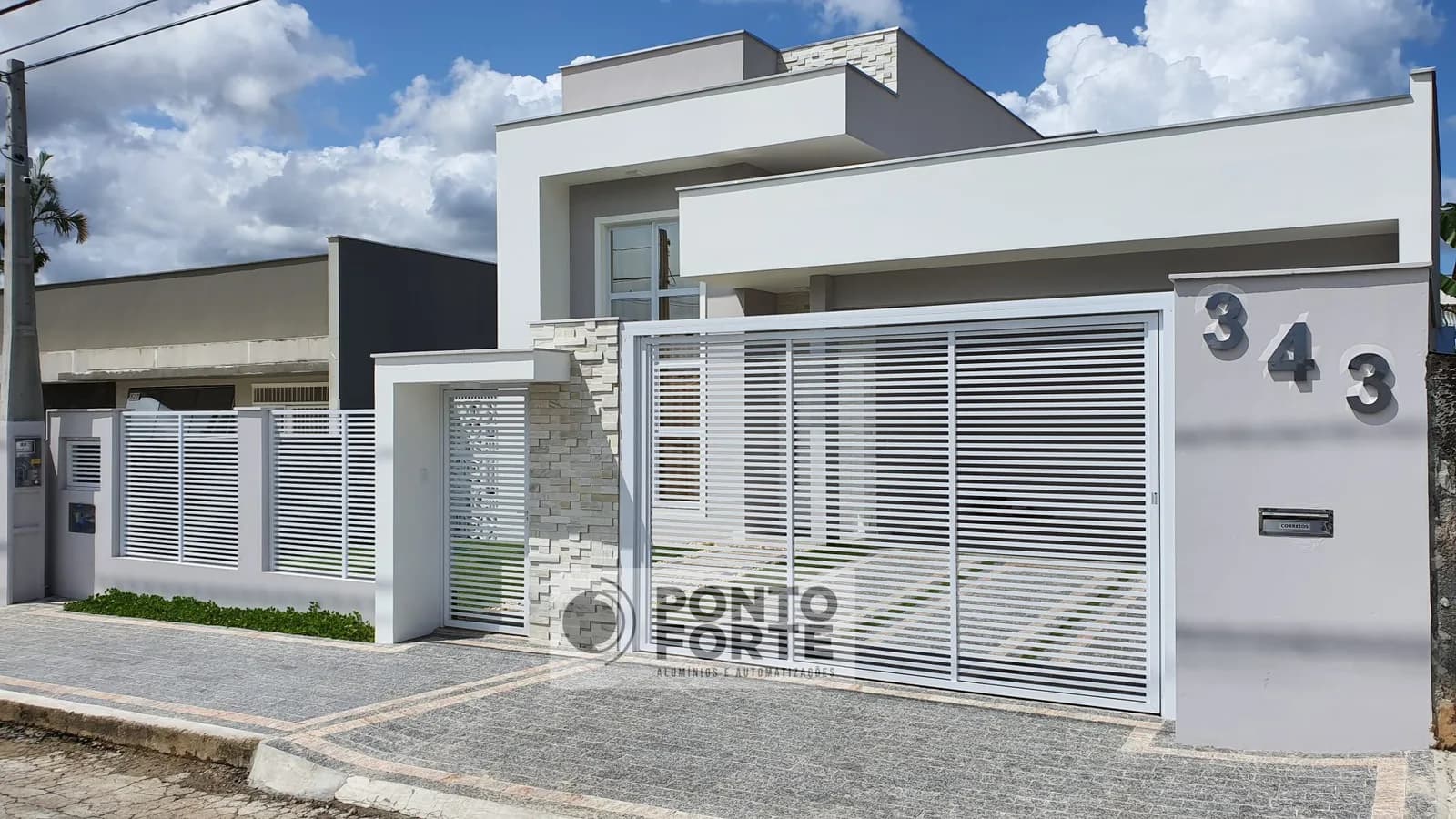 Portão basculante de alumínio branco vazado com barras, com portão social em vão próprio e cerca de alumínio no mesmo desenho, em residência de fachada clara em Joinville
