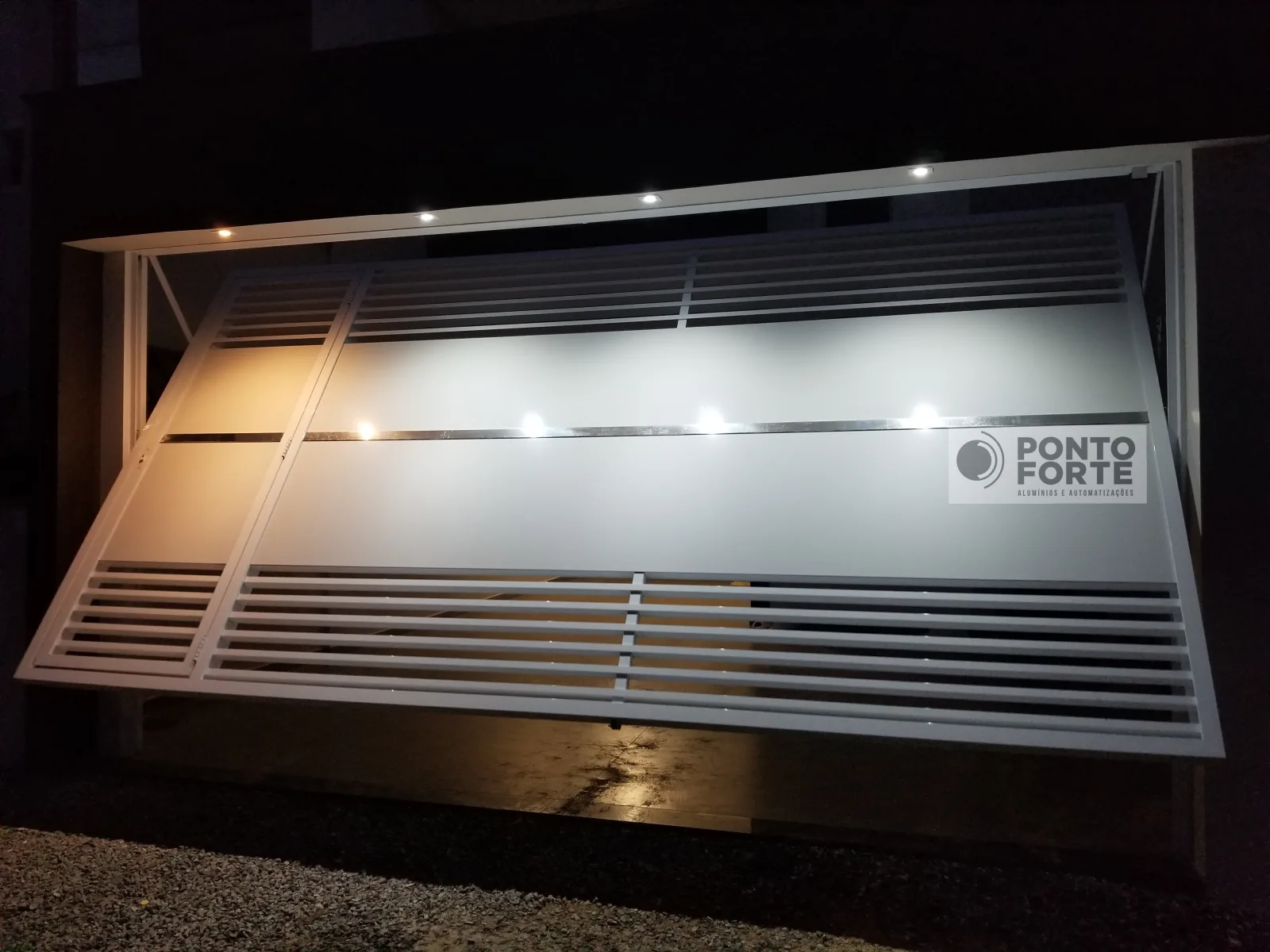Portão basculante de alumínio prata aberto, com a folha basculada para dentro da garagem iluminada, fechado no centro, com barras vazadas em cima e embaixo e portão social no mesmo quadro