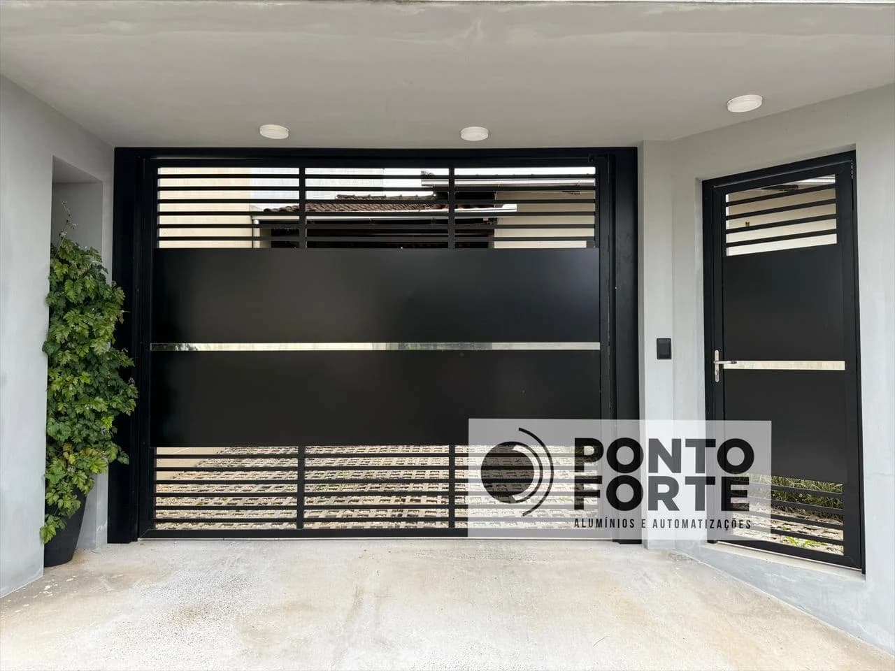 Portão basculante de alumínio preto fechado no centro, com faixa cromada e portão social ao lado em vão próprio, sob varanda de residência
