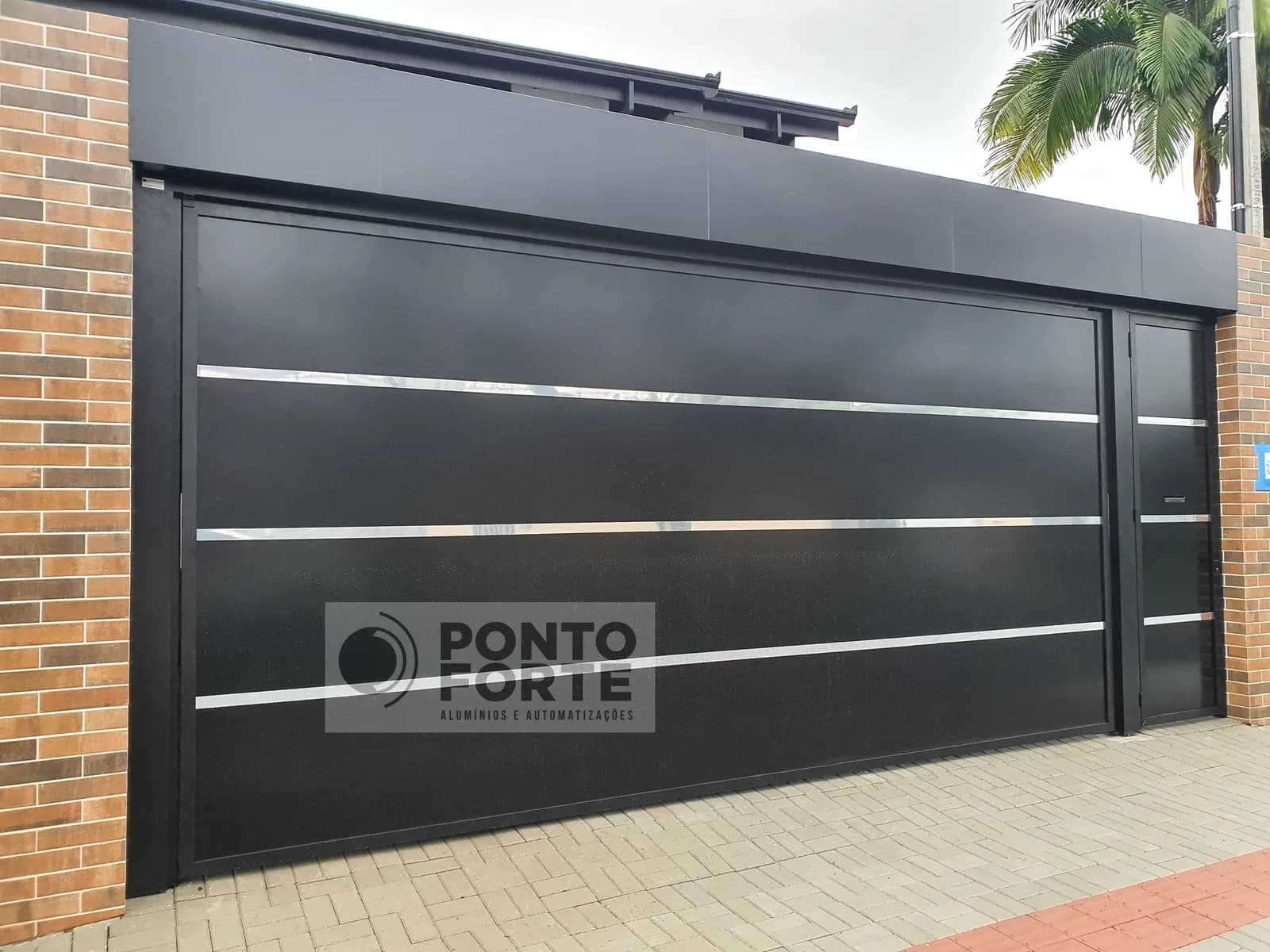 Portão basculante de alumínio preto em chapa lisa com faixas cromadas, feito sob medida, com o portão social em vão próprio no mesmo acabamento