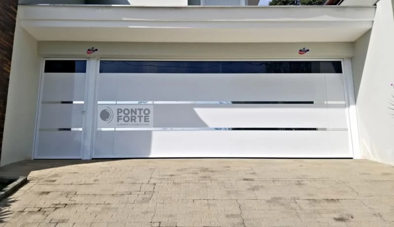 Portão de garagem largo de alumínio branco com faixas cromadas, faixa transparente no alto e porta social embutida no mesmo quadro, à esquerda